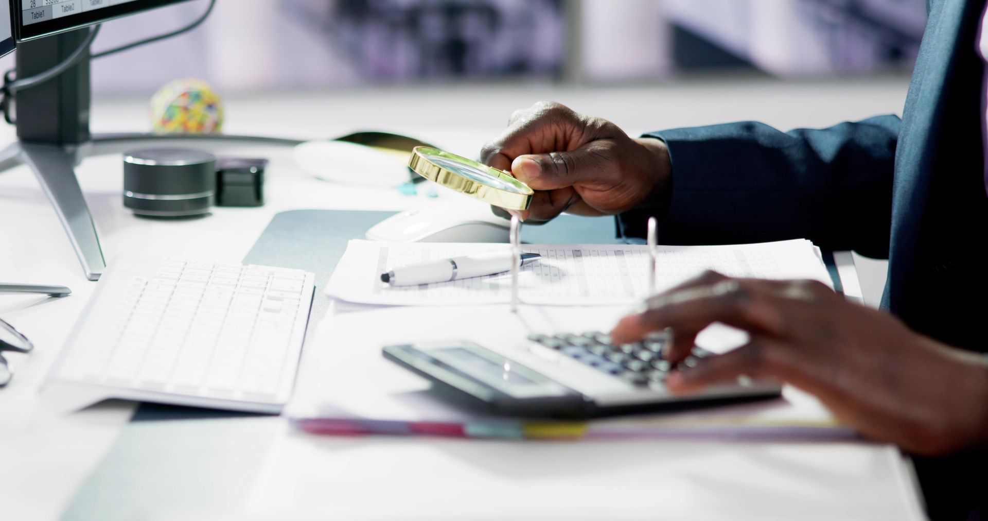 Person using a calculator and holding cash, working at a desk with papers, keyboard, and computer screen.
