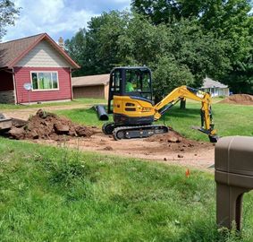 Yellow mini excavator digging dirt in a yard beside a red house and trees