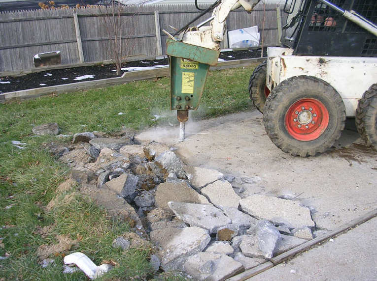 Bobcat with hydraulic breaker breaking cracked concrete driveway outdoors