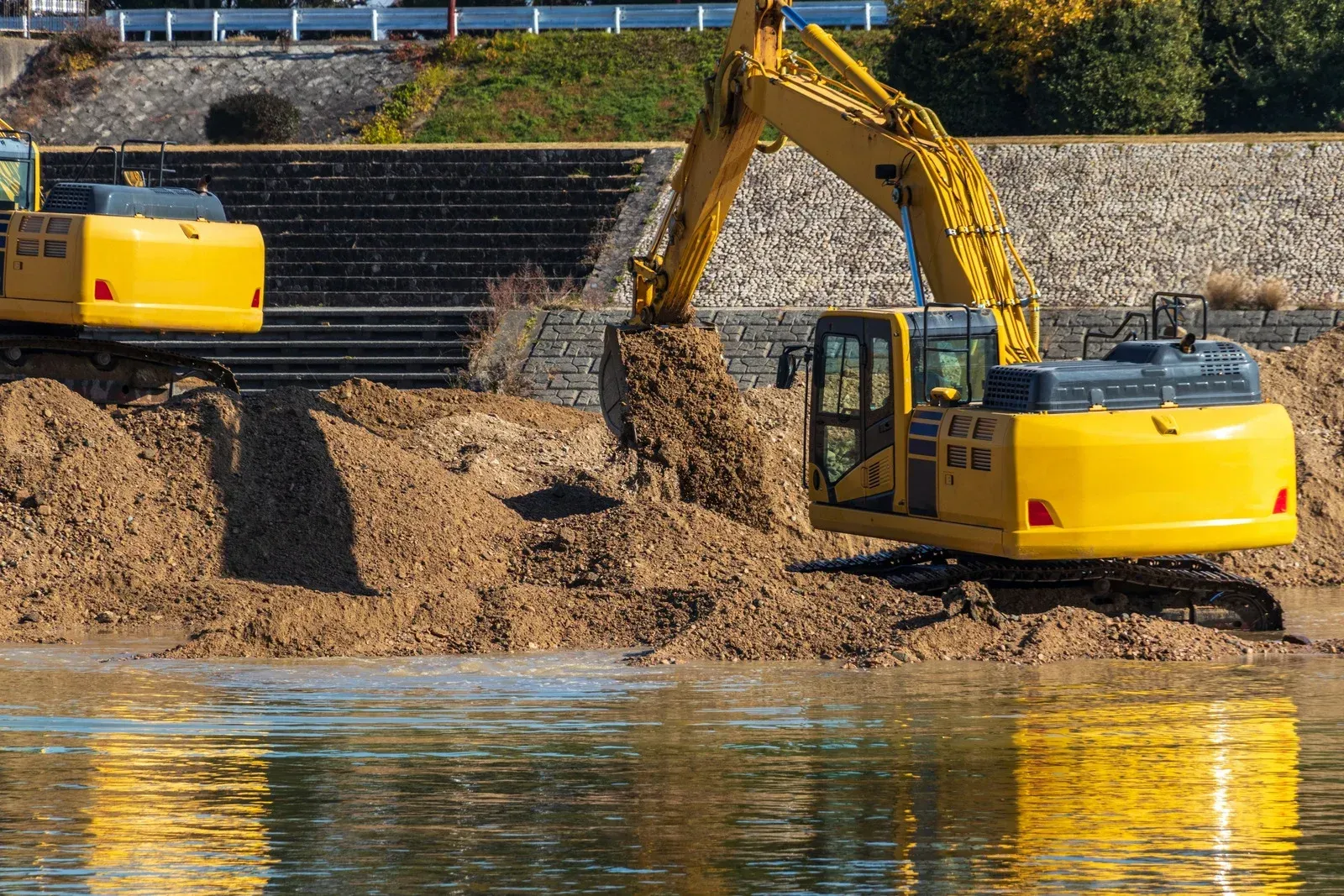 Two yellow excavators working on a sandy riverbank beside calm water