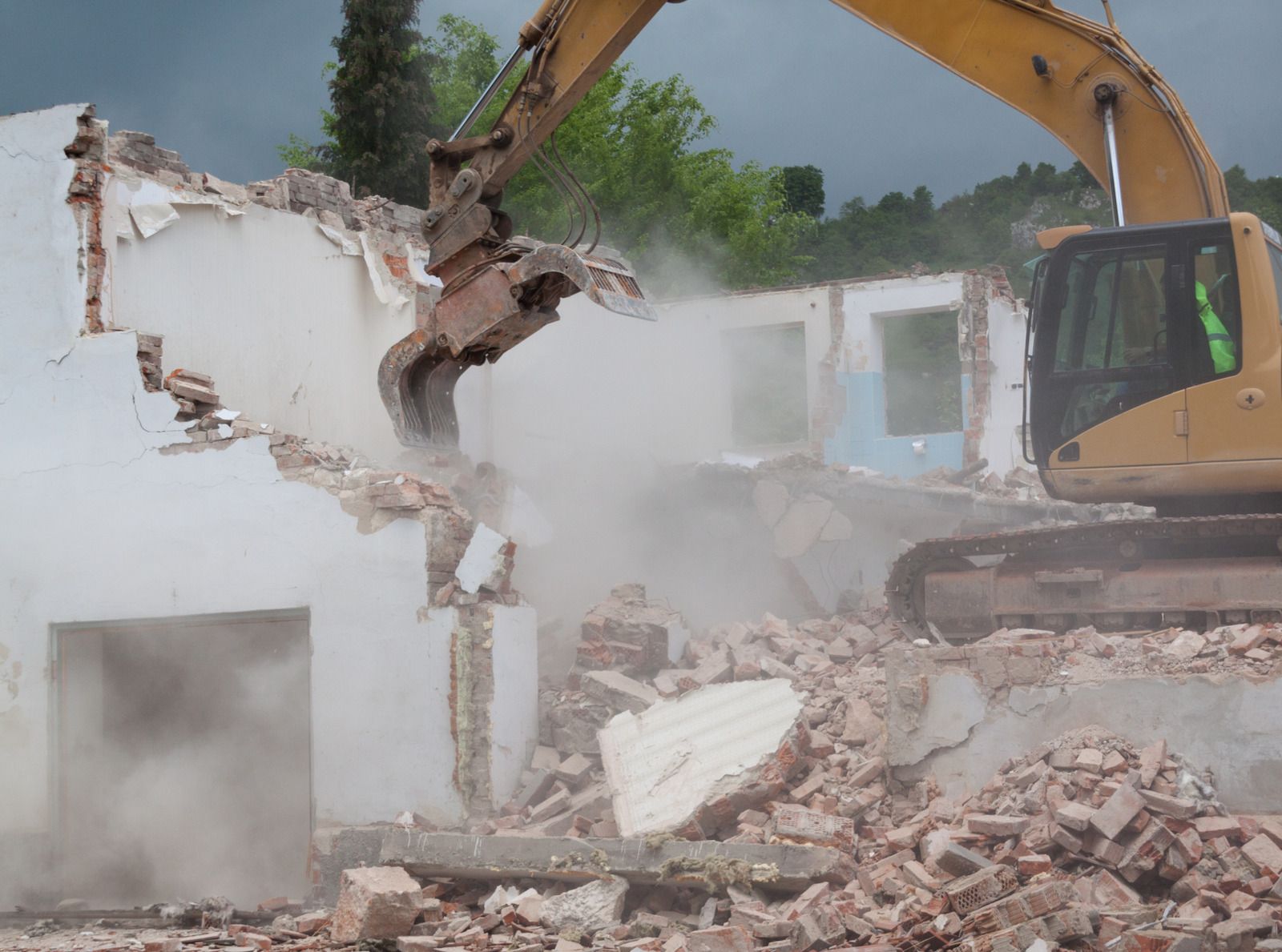 Excavator demolishing a white building, with dust and rubble filling the site.