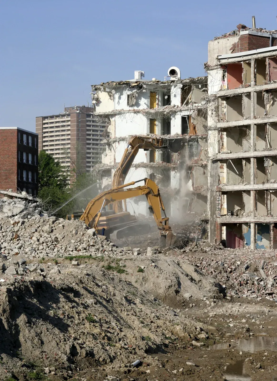 Yellow excavator demolishing rubble beside damaged buildings under a clear blue sky