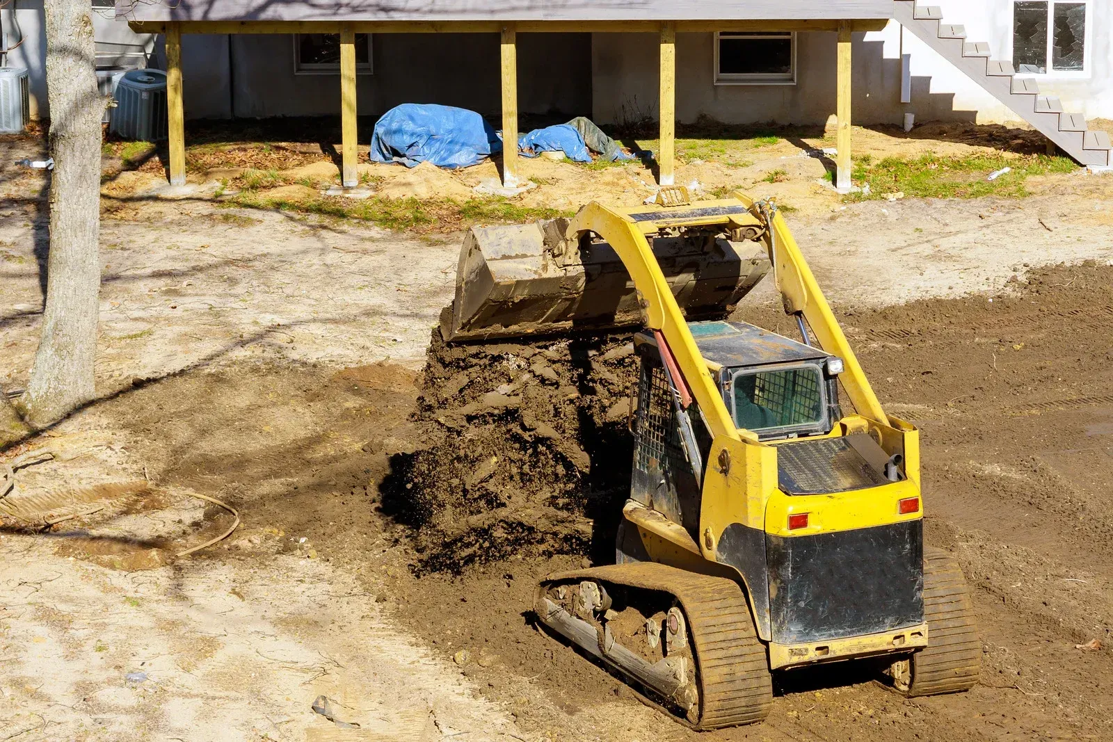Small excavator dumping dirt in a muddy construction area near a house