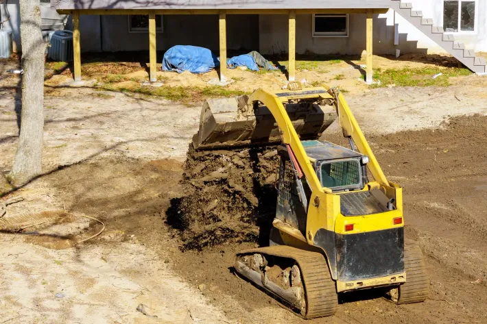 Small excavator dumping dirt in a muddy construction area near a house