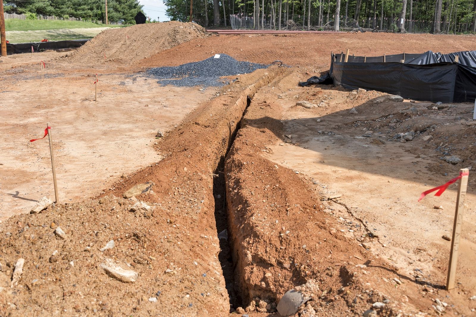 Construction site with a deep trench cut through brown dirt, marked by red flags and black silt fencing.