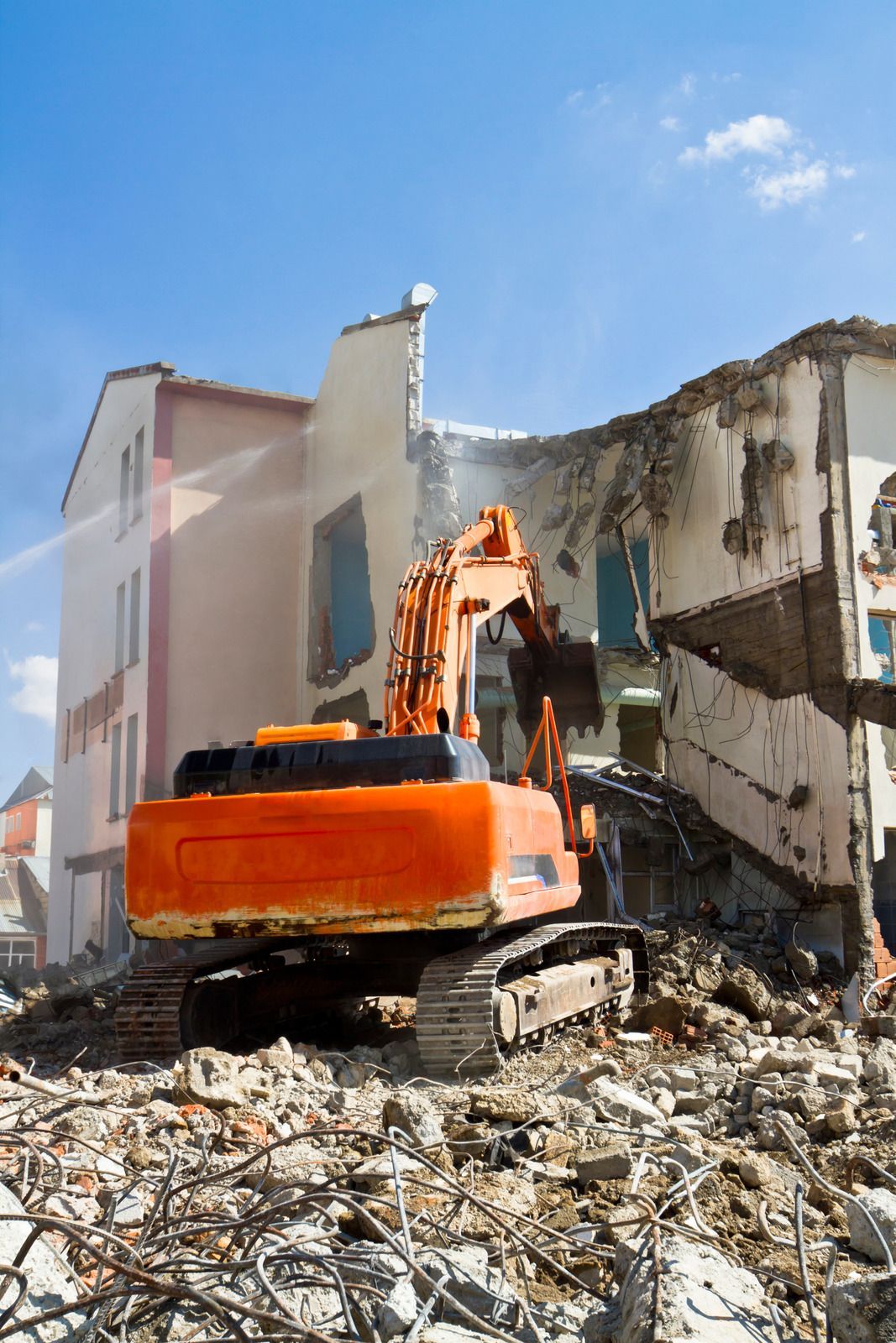 Orange excavator demolishing a partially collapsed building amid rubble under a blue sky