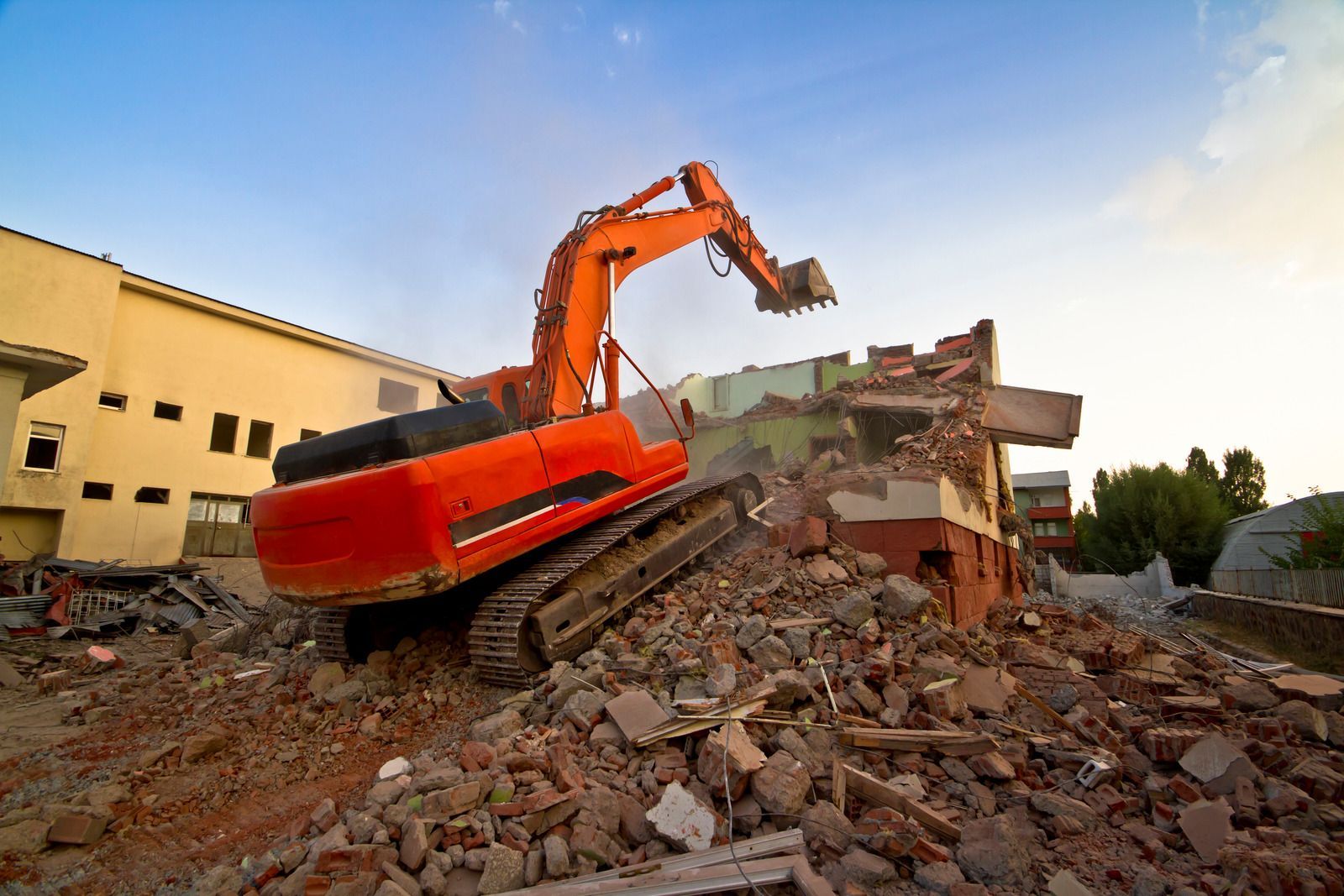 Orange excavator demolishing a rubble pile beside a building at sunset
