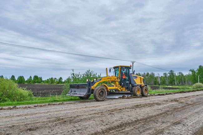 Yellow road grader on a muddy rural road under a cloudy sky