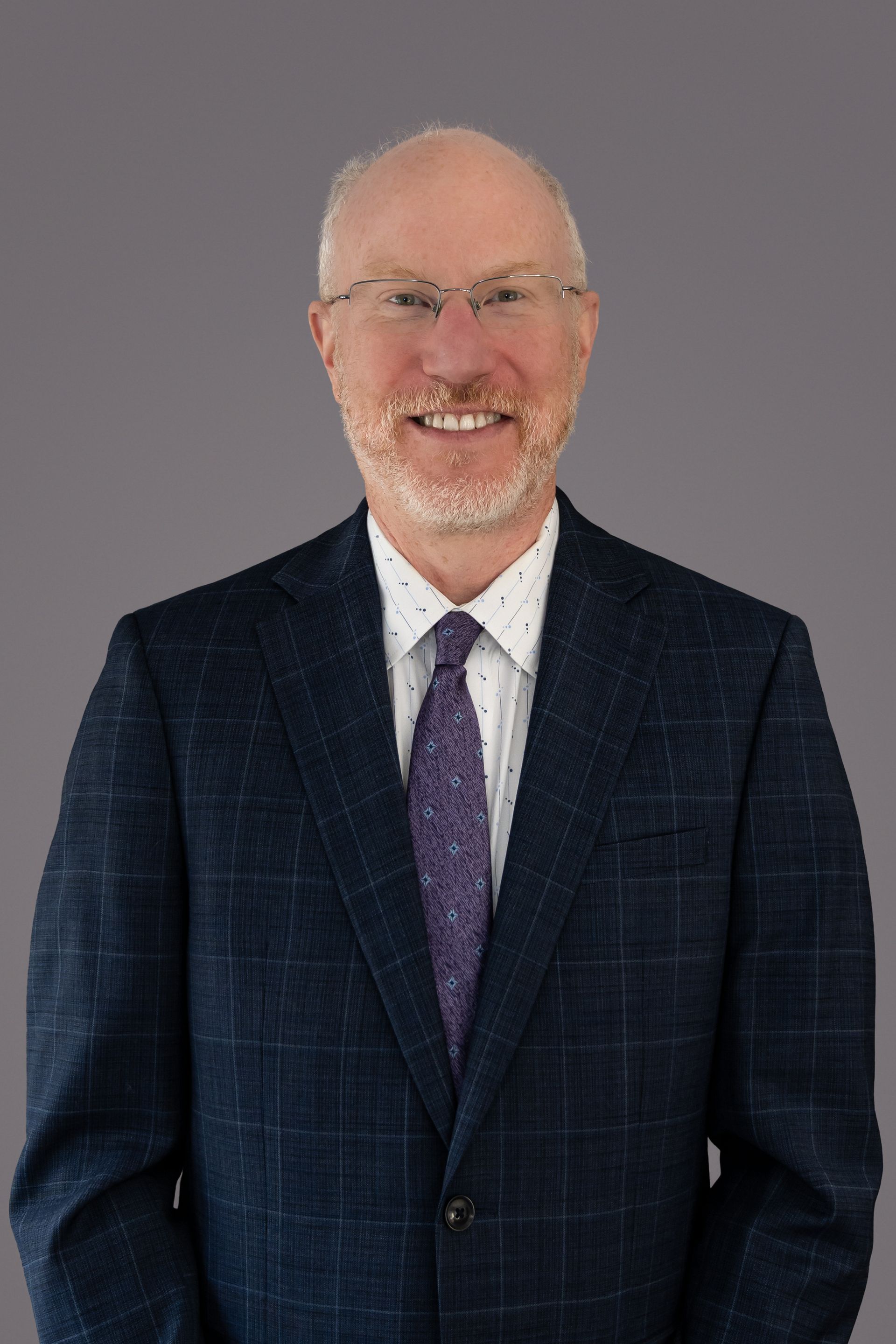 Robery G Kayser Man in a suit smiles at the camera, with glasses and a patterned tie, set against a grey background.