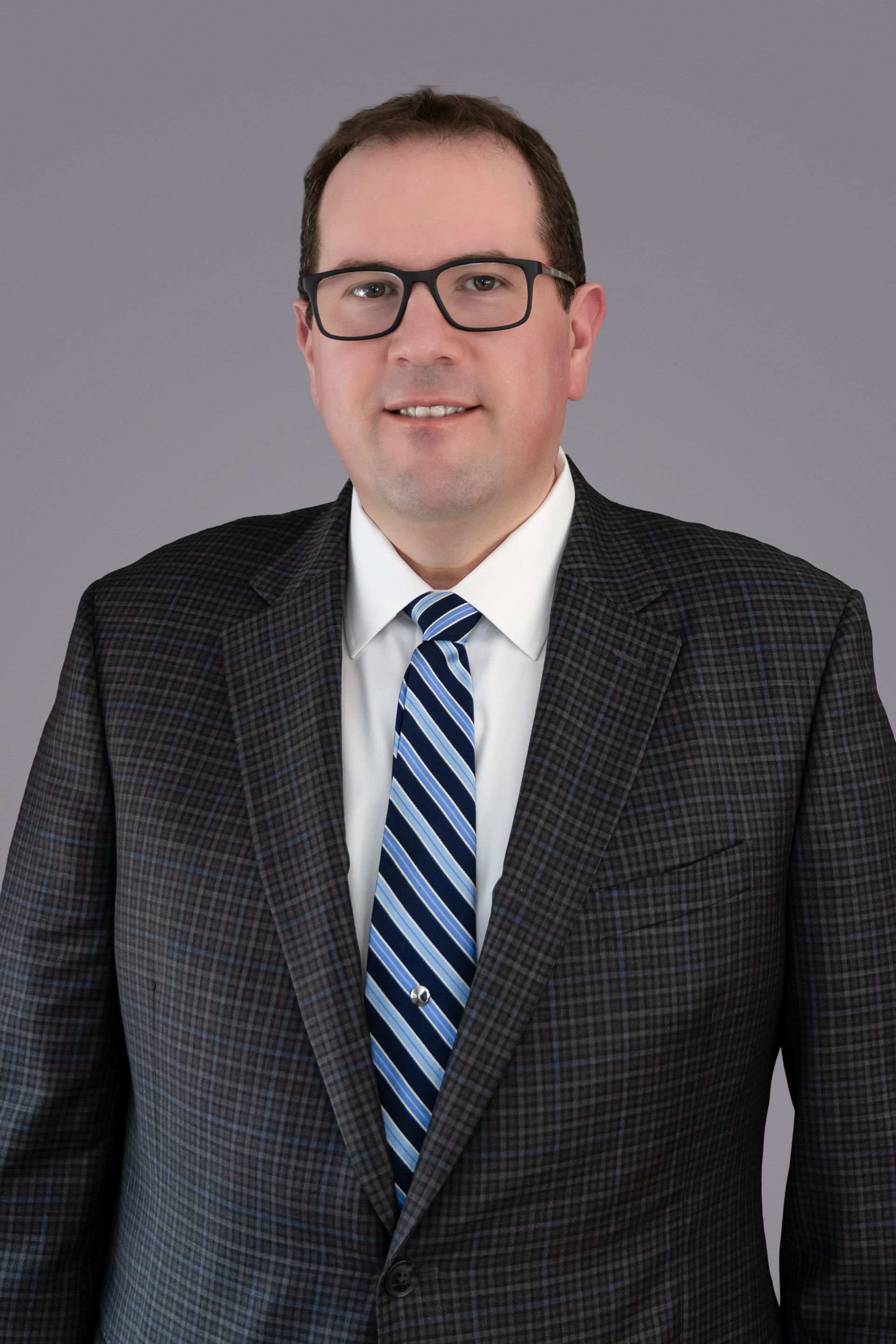 Man wearing glasses, suit, and tie smiling, against a gray backdrop.