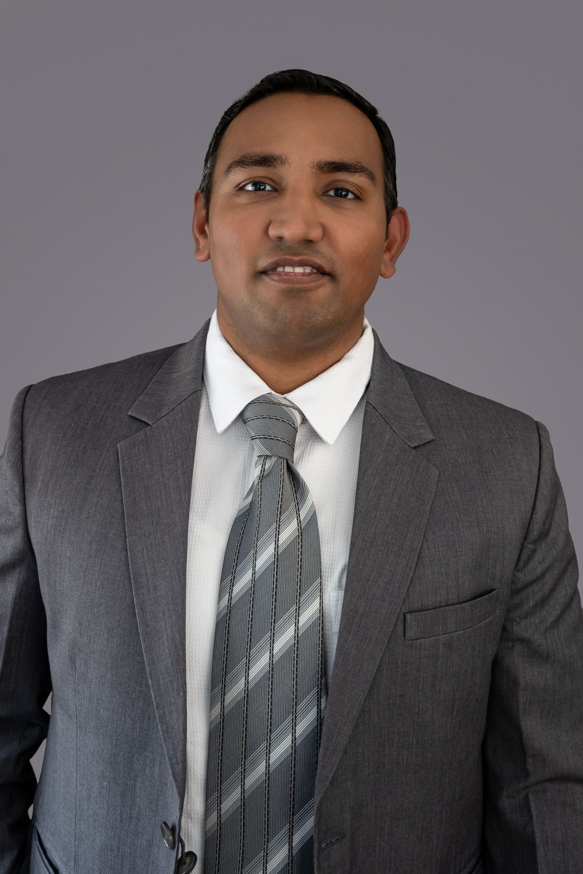 Man in gray suit and patterned tie against a gray background, smiling slightly.