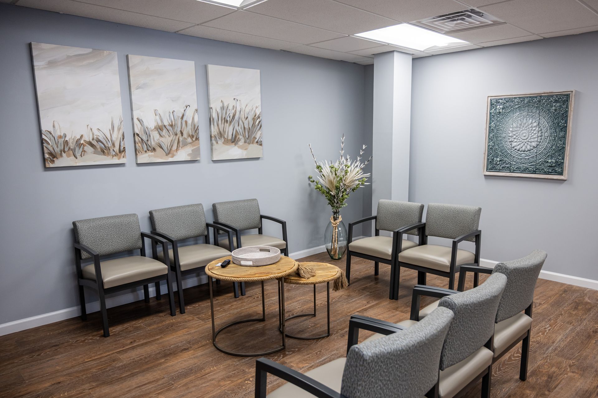 Waiting room with chairs, art, and small tables. Gray walls, light wood floor.