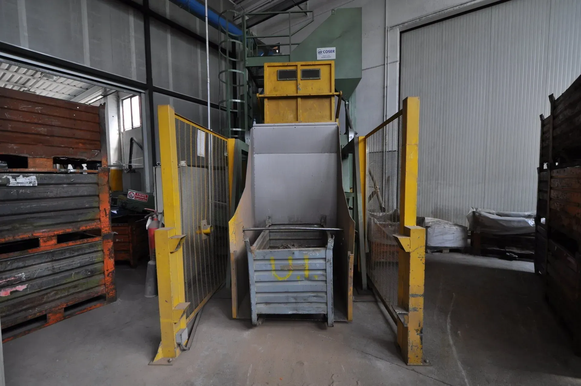 Compattatore di rifiuti industriali in un magazzino, con porte di sicurezza gialle e bidoni impilati davanti.
