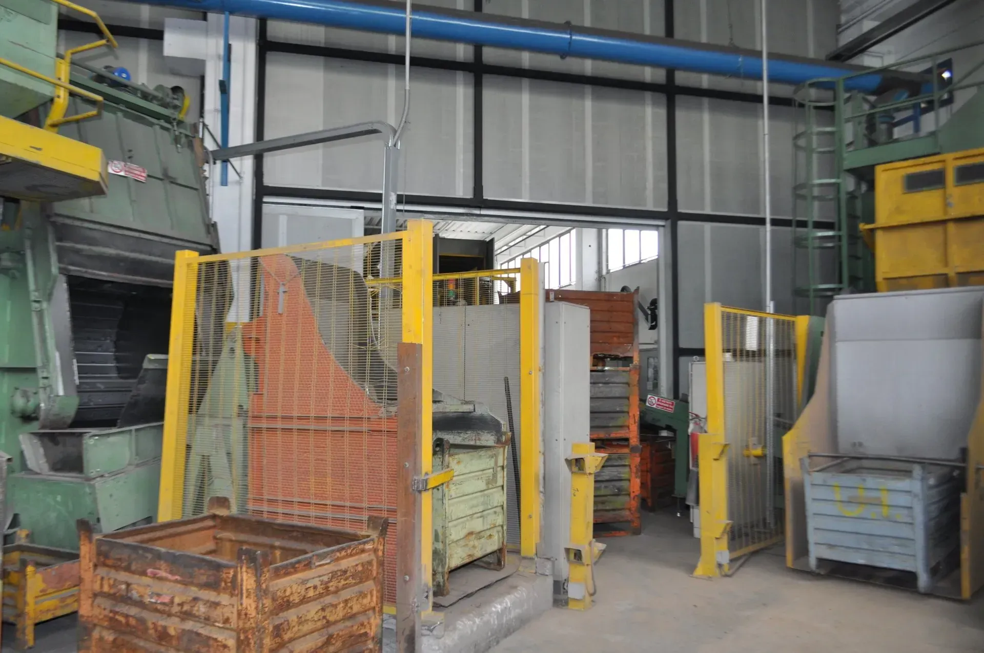 Magazzino industriale con cancelli di sicurezza gialli, macchinari metallici e materiali impilati all'interno di un'area di lavoro di fabbrica.