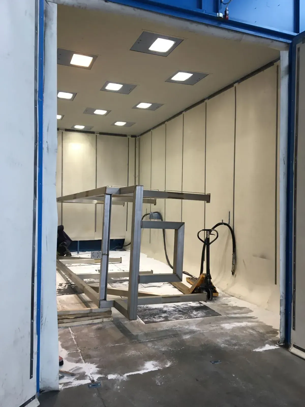 Locale industriale vuoto con porta blu, luci a soffitto e un tavolo da lavoro in metallo al centro.
