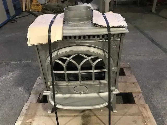 In un magazzino, un inserto metallico per stufa è fissato a un pallet, con tubi impilati e imbottitura in schiuma sopra.