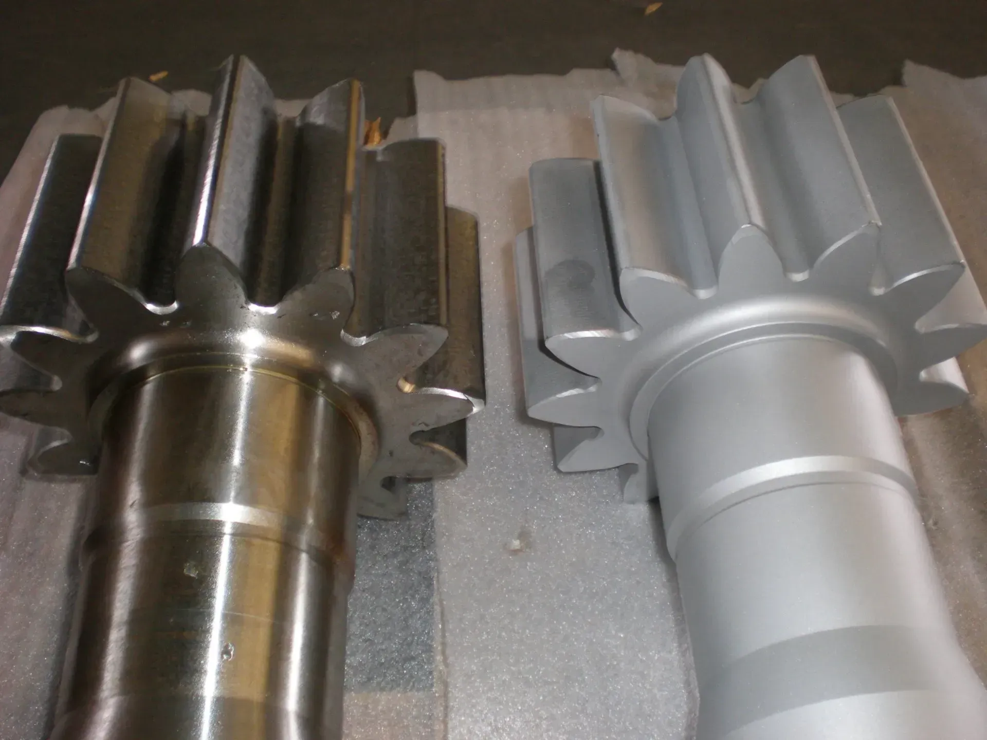 Due alberi di ingranaggi metallici, uno in acciaio scuro e uno argentato, giacciono uno accanto all'altro su una superficie.