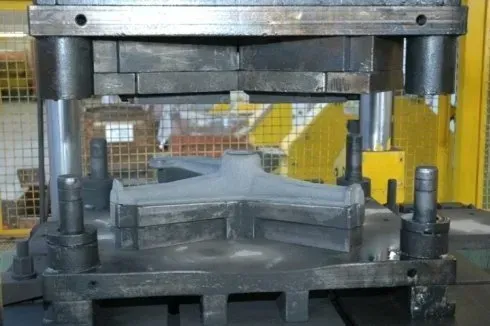 Una pressa industriale modella un componente metallico in una fabbrica, con macchinari gialli sullo sfondo.