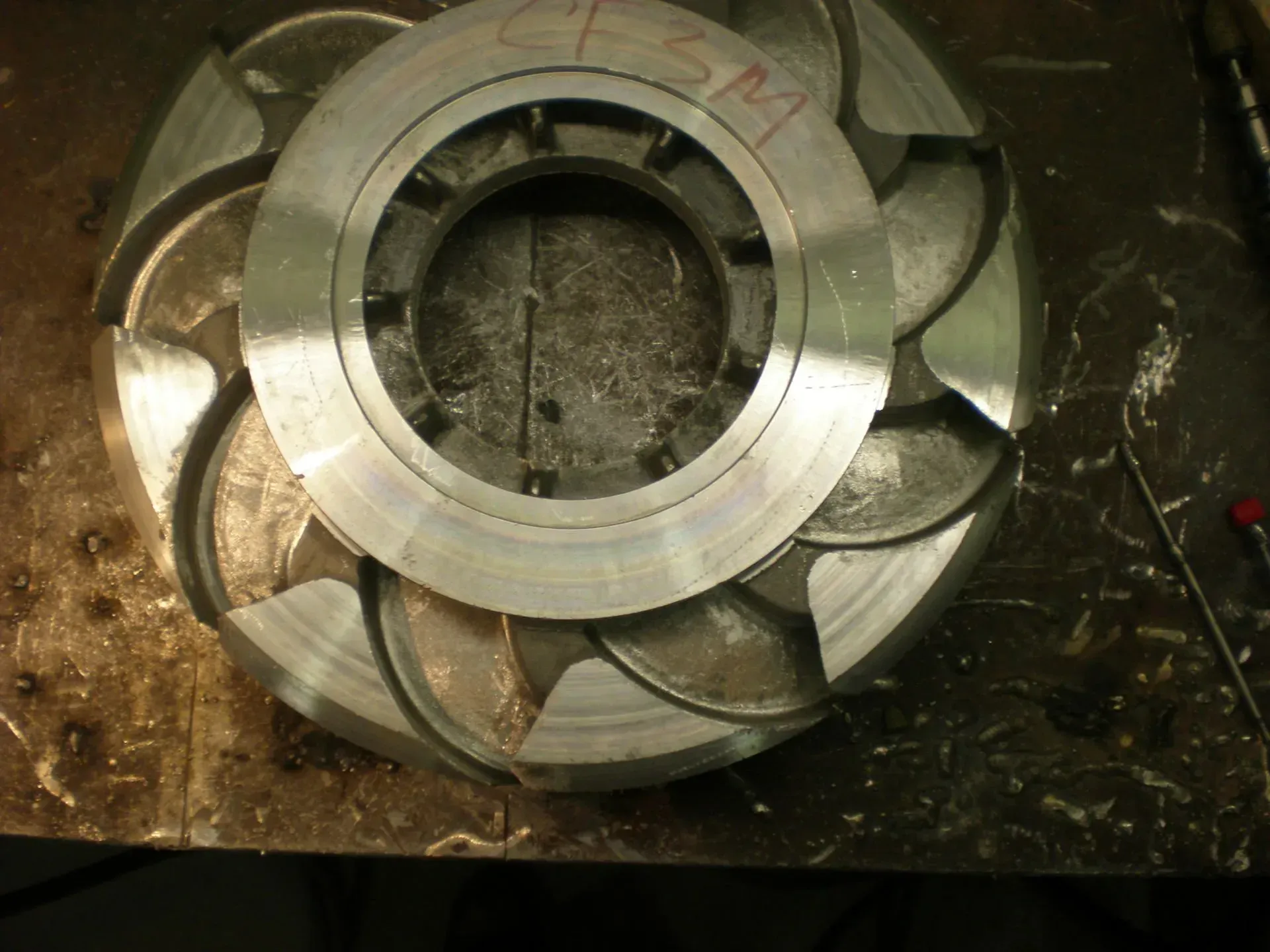 Pezzo circolare in metallo, simile a un ingranaggio, con lame curve, lavorato su una superficie di officina.