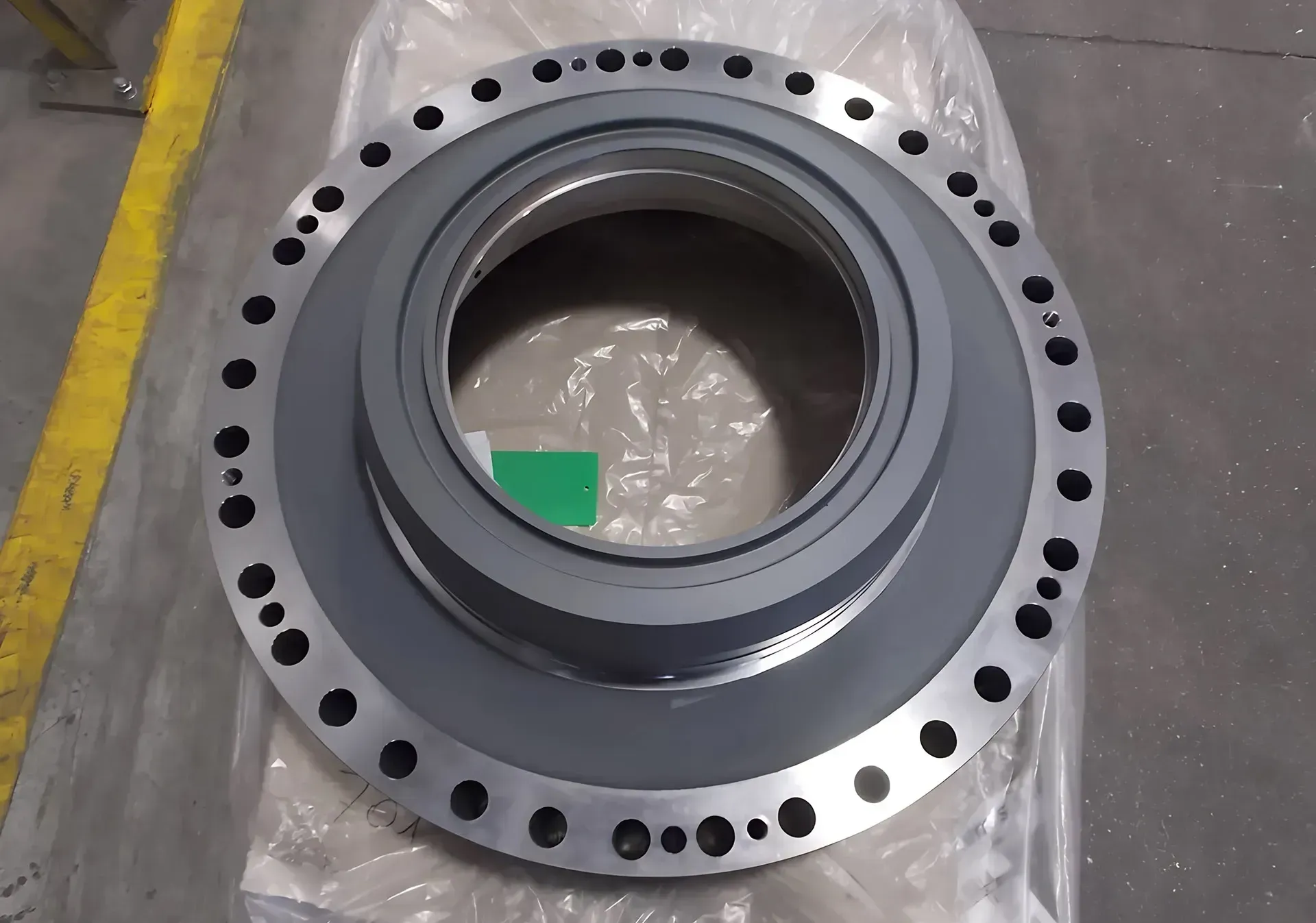 Flangia ad anello in metallo con fori per bulloni equidistanti, appoggiata su pellicola di plastica sul pavimento di un'officina.