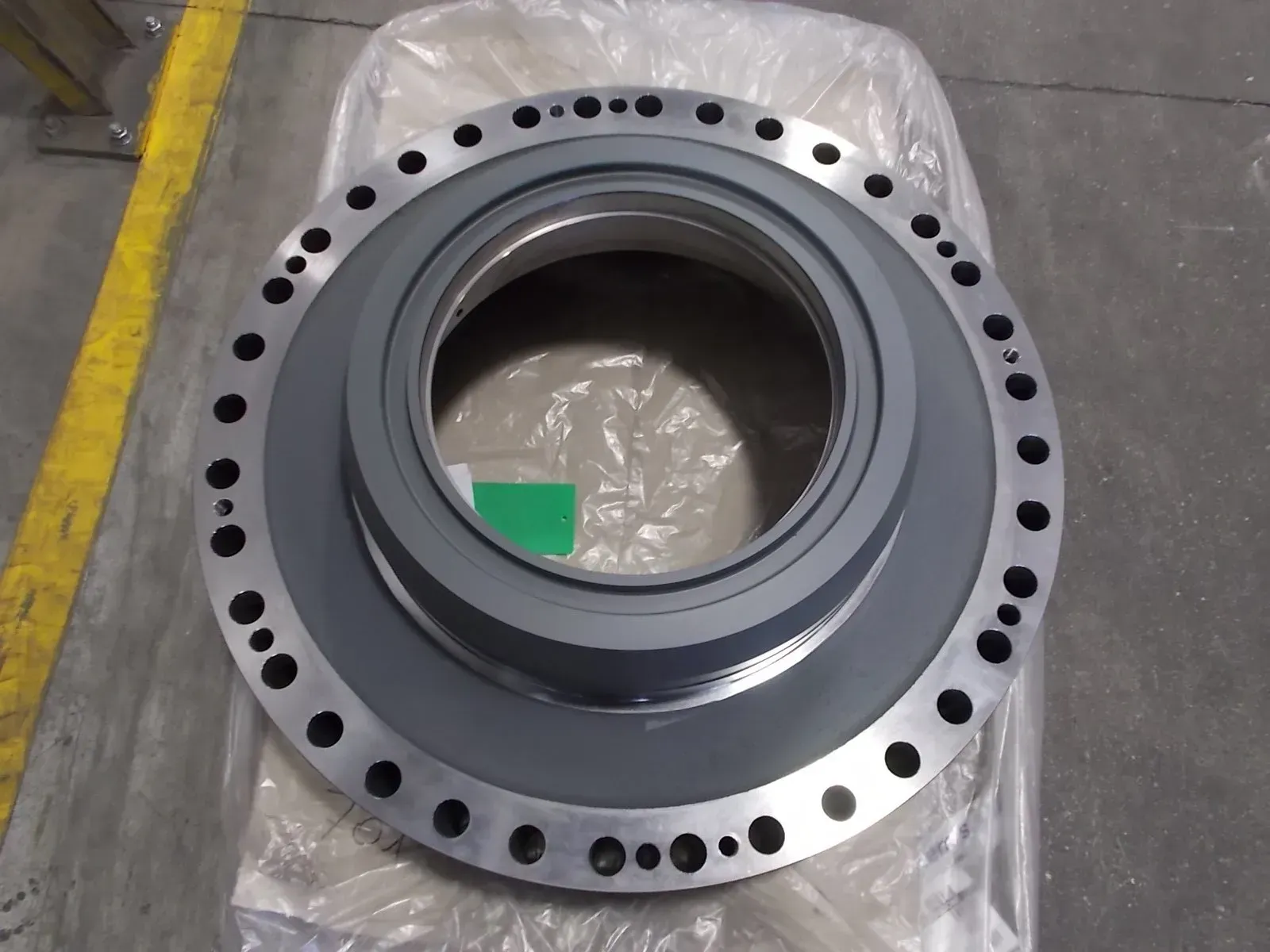 Grande anello metallico grigio con fori per bulloni, avvolto nella plastica, sul pavimento di un magazzino.