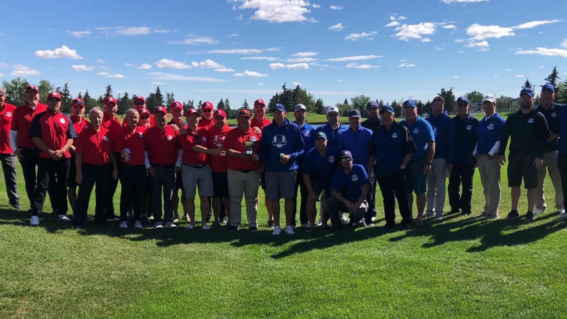 Groups of people in red and blue shirts stand on a green lawn with a golf course in the background.
