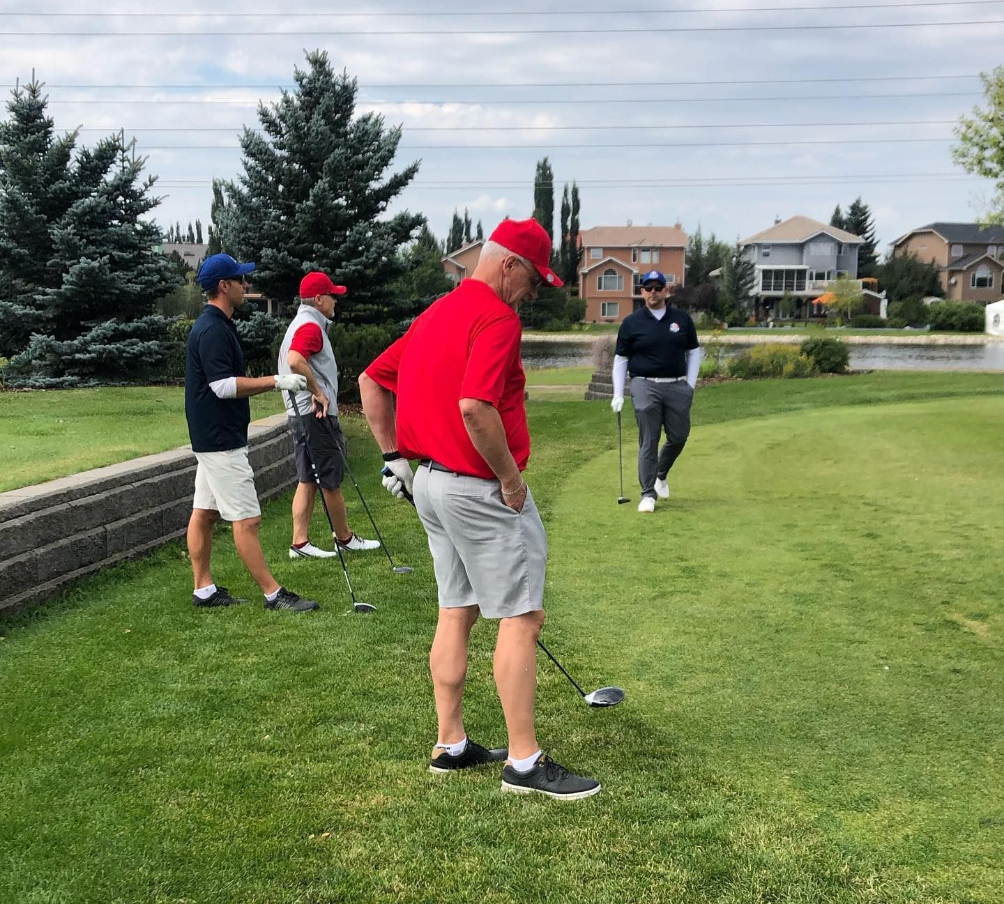Golfers on a green. Man in red shirt prepares to hit; others watch, holding clubs. Sunny day.