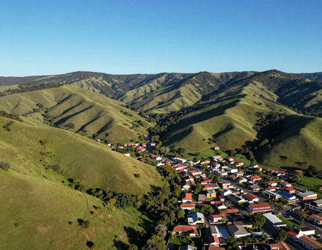 Vista aérea de un pequeño pueblo en un valle verde rodeado de colinas onduladas bajo un cielo azul claro.