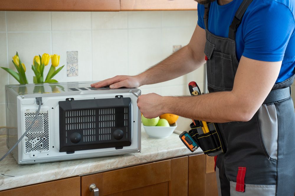 Person repairing a microwave on a kitchen counter, with tools and yellow tulips nearby.