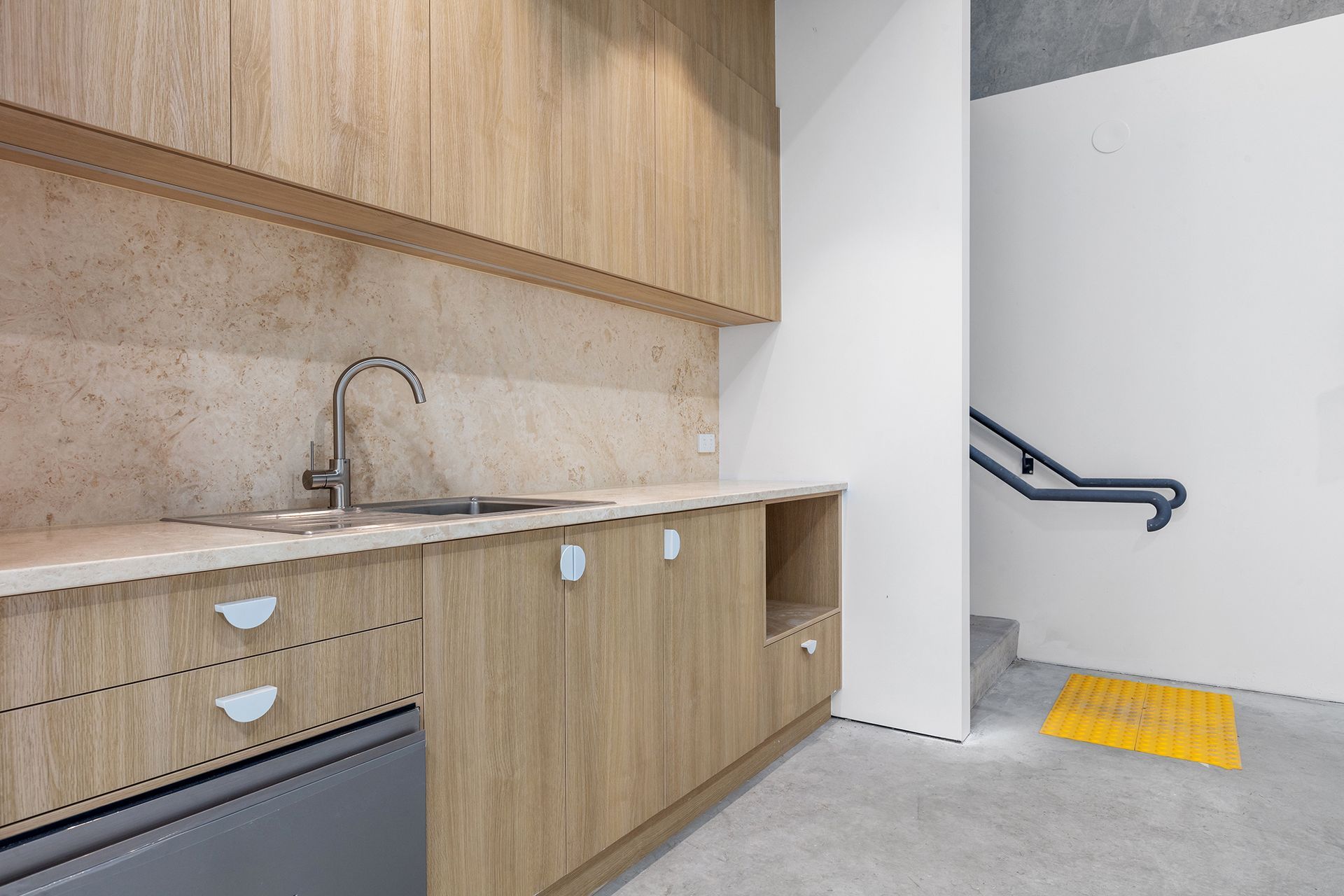 A light-wood kitchenette with a metal sink beside a white wall with a handrail and a yellow tactile paving tile on the floor.