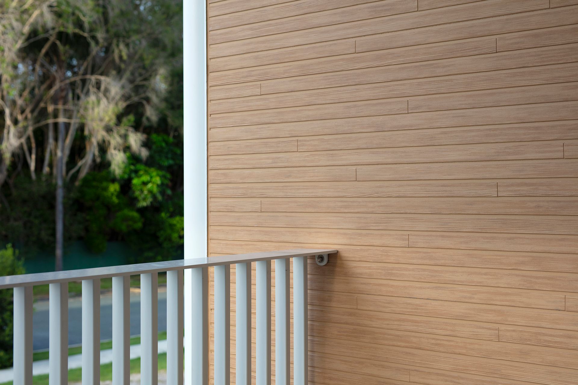 A close-up view of a light-brown, horizontal wood-panel wall beside a white metal railing overlooking a blurry, green scene.