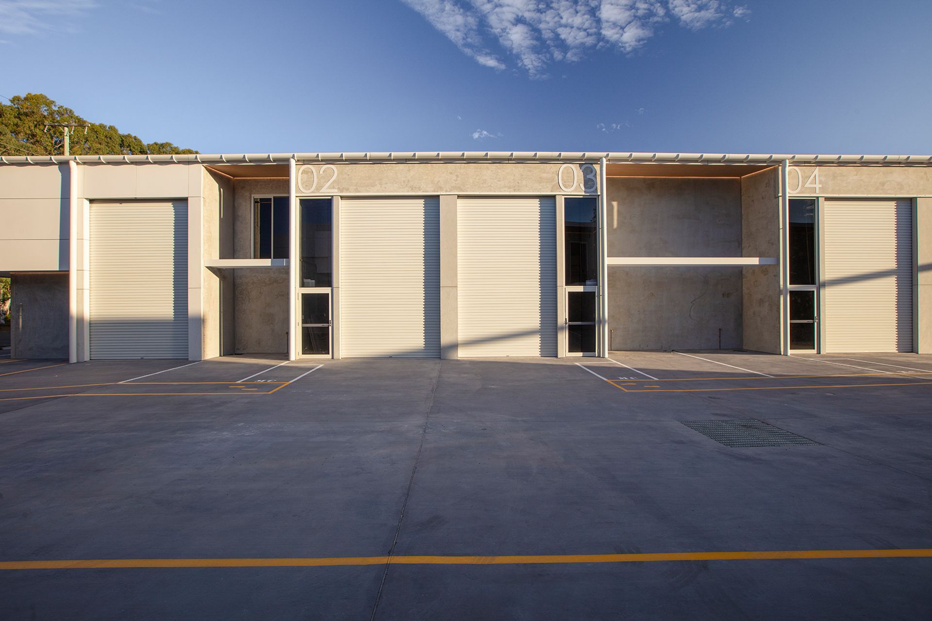 A row of light-colored, modern industrial warehouse units with roll-up doors and vertical windows under a blue sky.