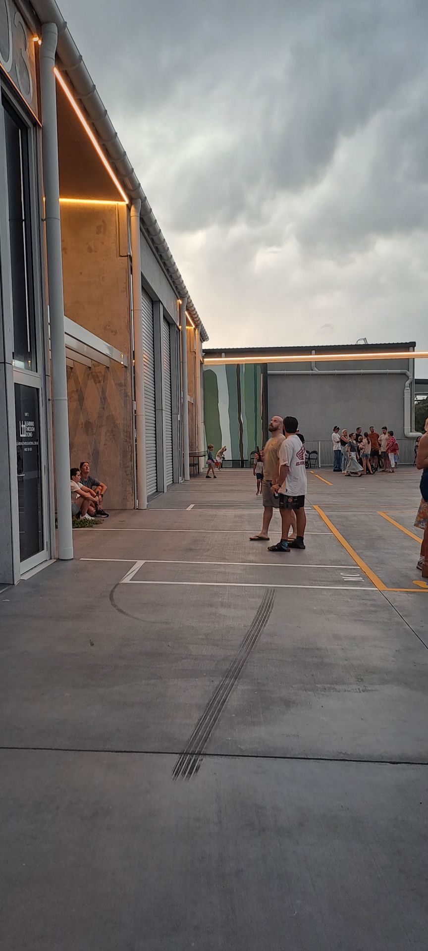 People stand in a paved outdoor plaza beside a modern building with warm interior lighting under a cloudy sky.