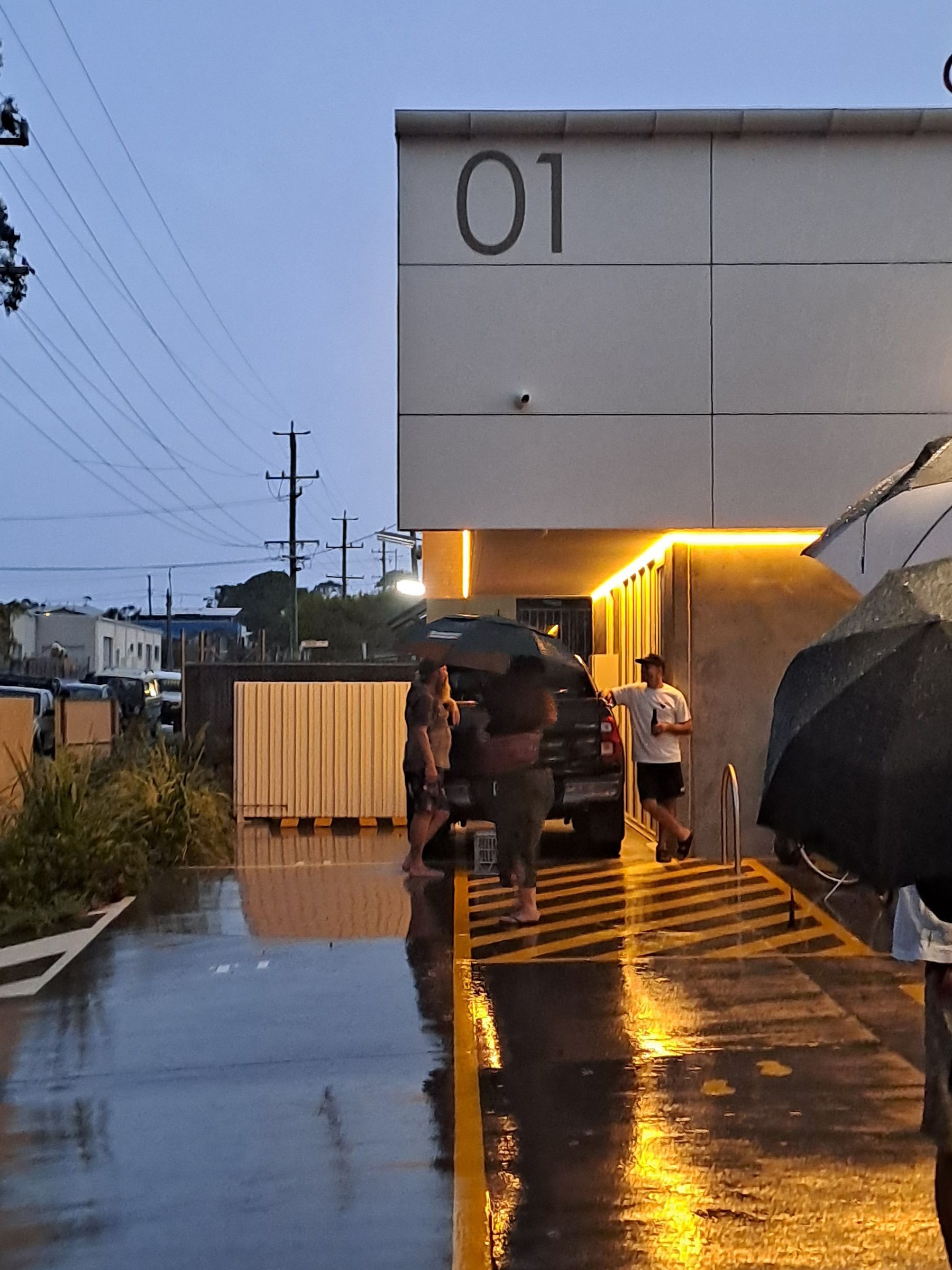 A small group stands near a vehicle parked under building 01 at dusk, with wet ground reflecting warm building lights.