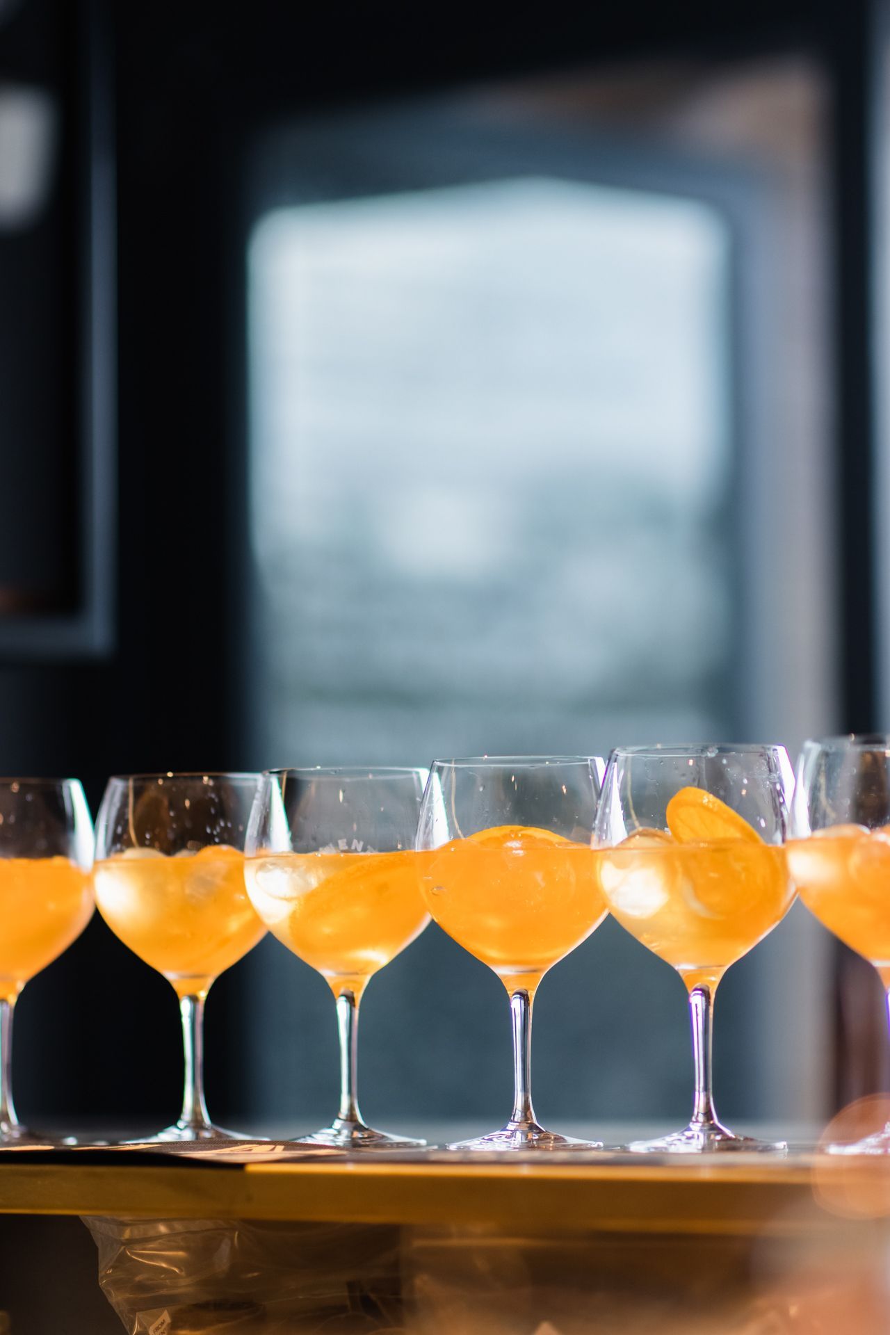 A row of six stemmed glasses filled with an orange cocktail and ice sits on a bar counter in a dimly lit room.