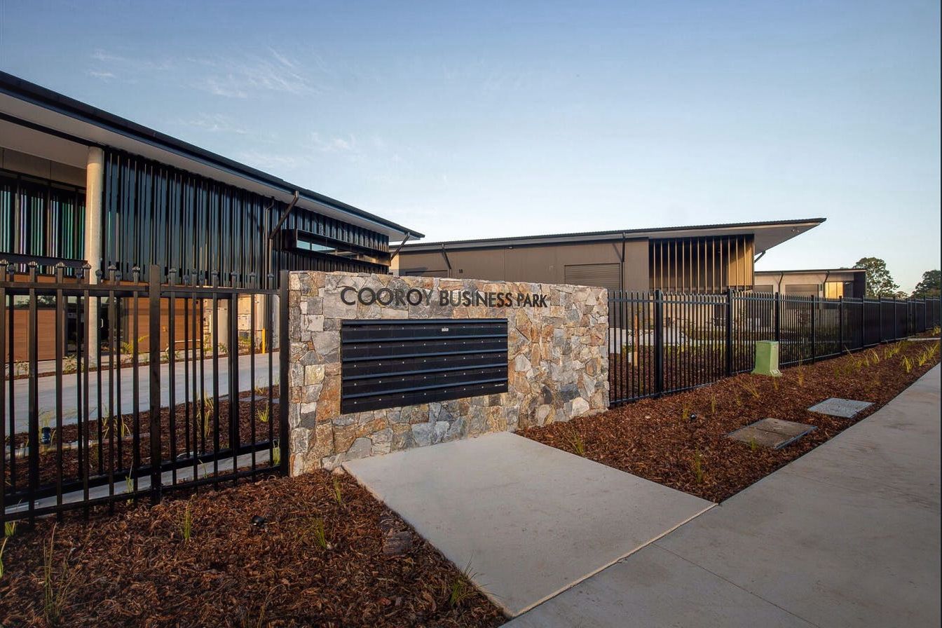 A low stone wall with a black mailbox bank marks the entrance to the Cooroy Business Park in front of modern buildings.