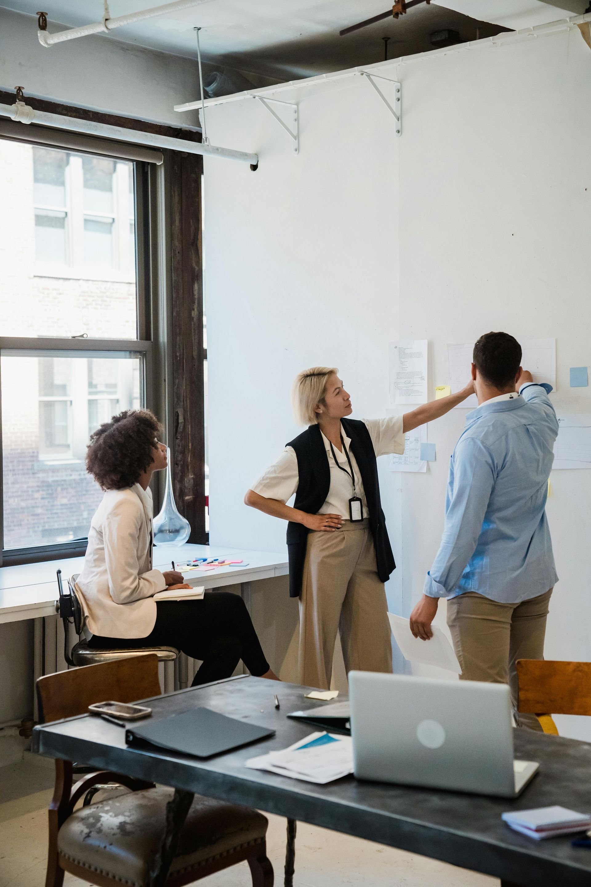 Three colleagues collaborate in a bright office, with two standing by a wall of notes and one seated at a desk.