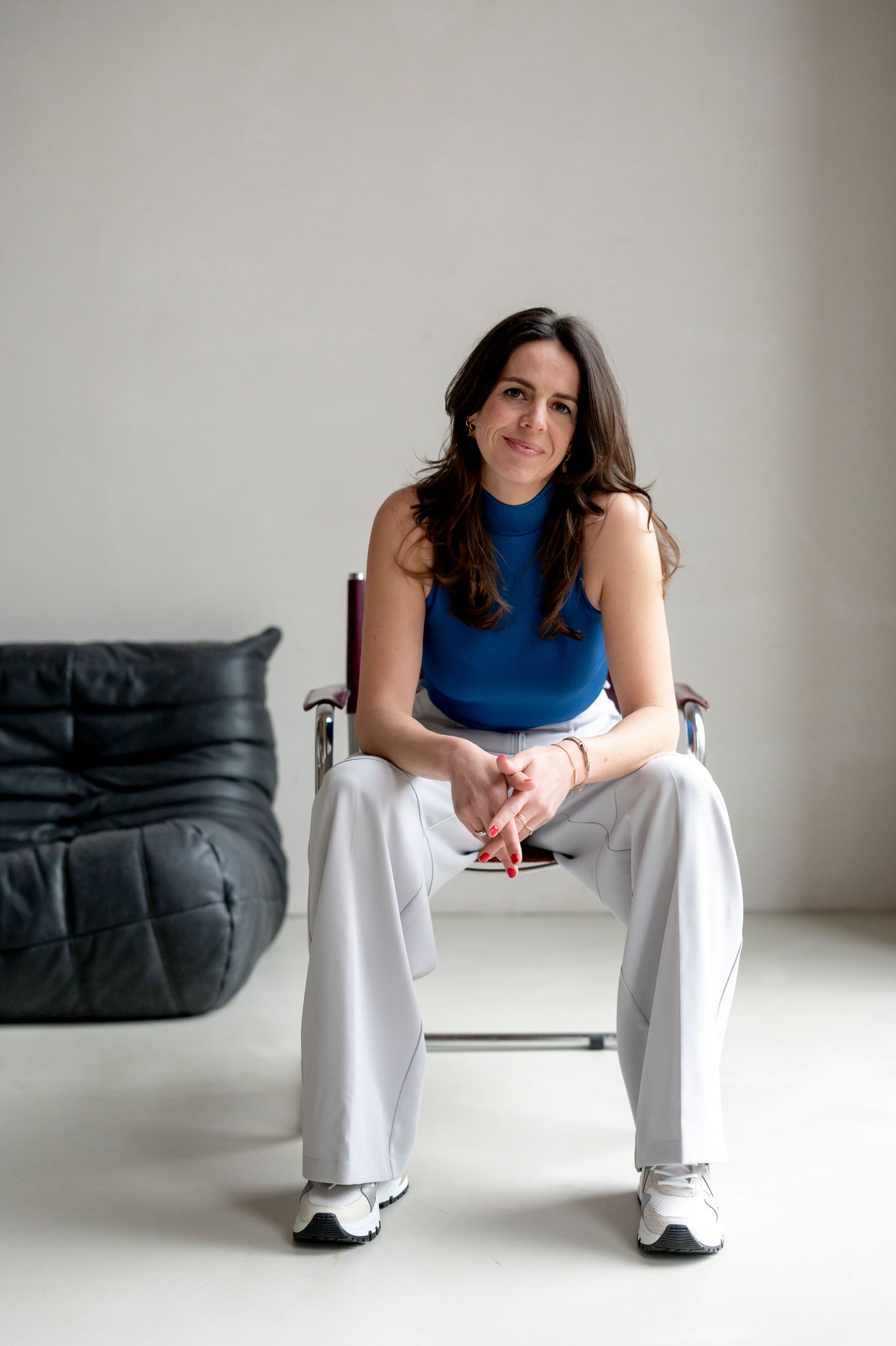 Een vrouw in een blauwe top en witte broek zit glimlachend op een stoel.