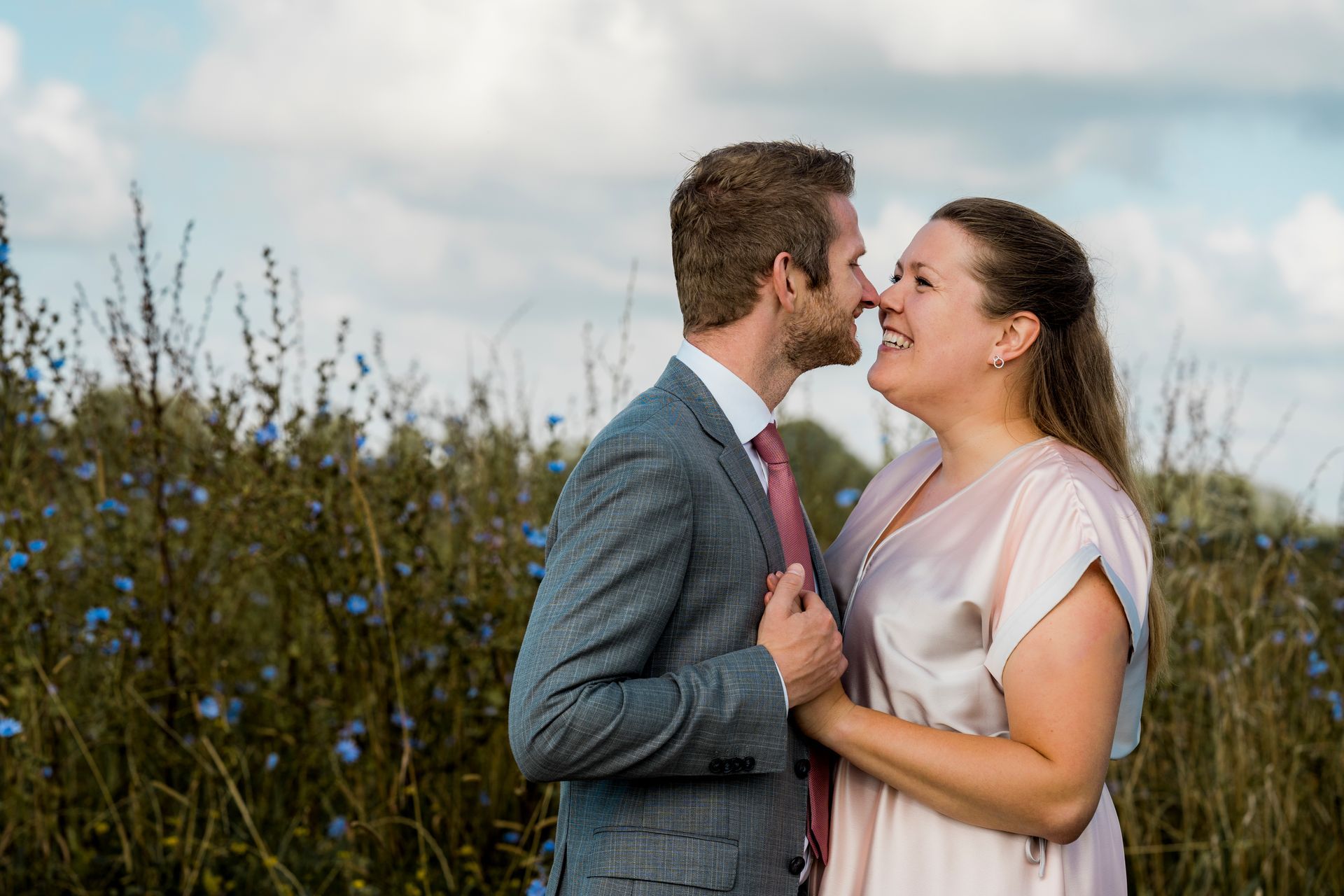 Een stel omhelst elkaar in de buitenlucht; de man kust de vrouw op haar neus. Ze glimlachen, omringd door blauwe bloemen onder een bewolkte hemel.