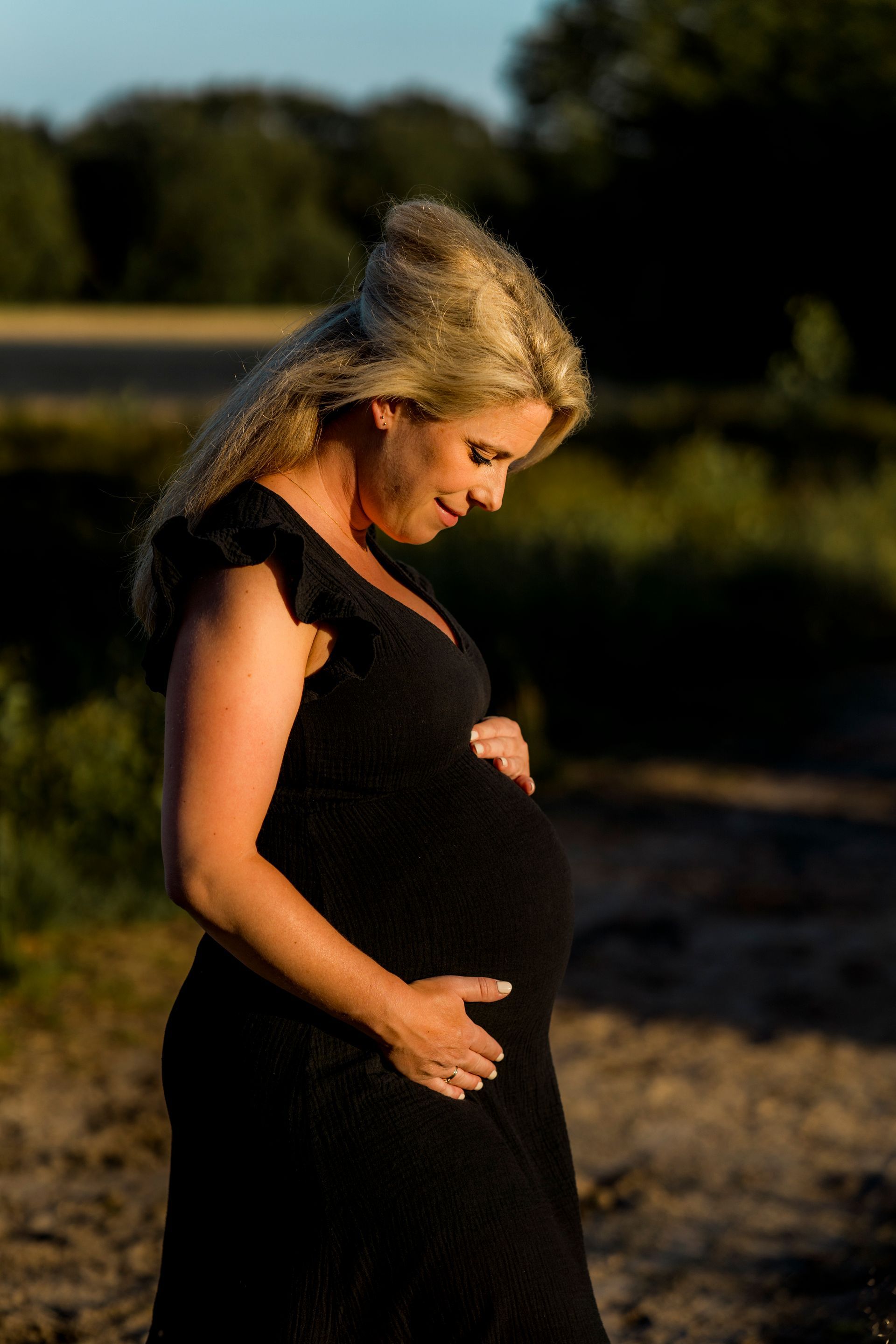 Zwangere vrouw in een zwarte jurk, die zachtjes haar buik aanraakt. Buiten, in een veld, glimlachend.