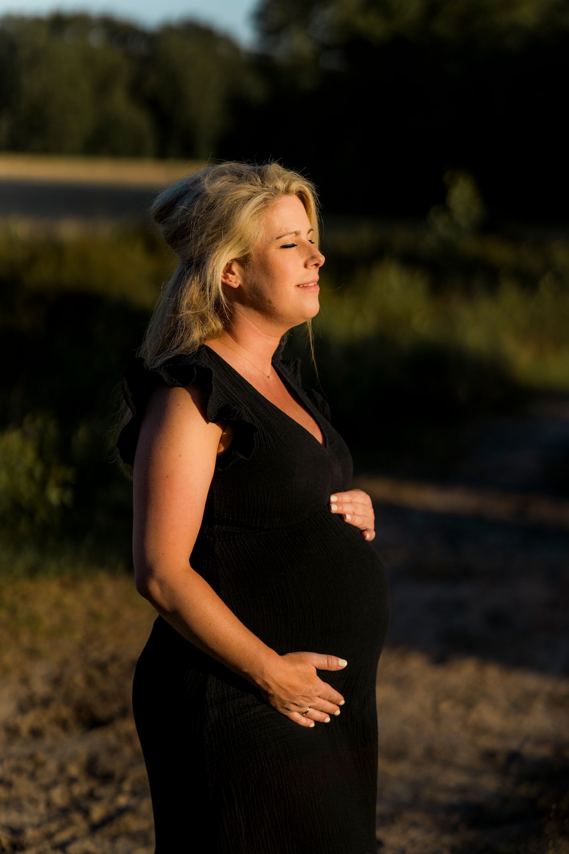 Zwangere vrouw in zwarte jurk, buiten, houdt haar buik vast, ogen gesloten, zonlicht.