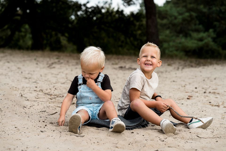 Twee blonde kinderen zitten in het zand, de ene lacht, de andere kijkt peinzend.