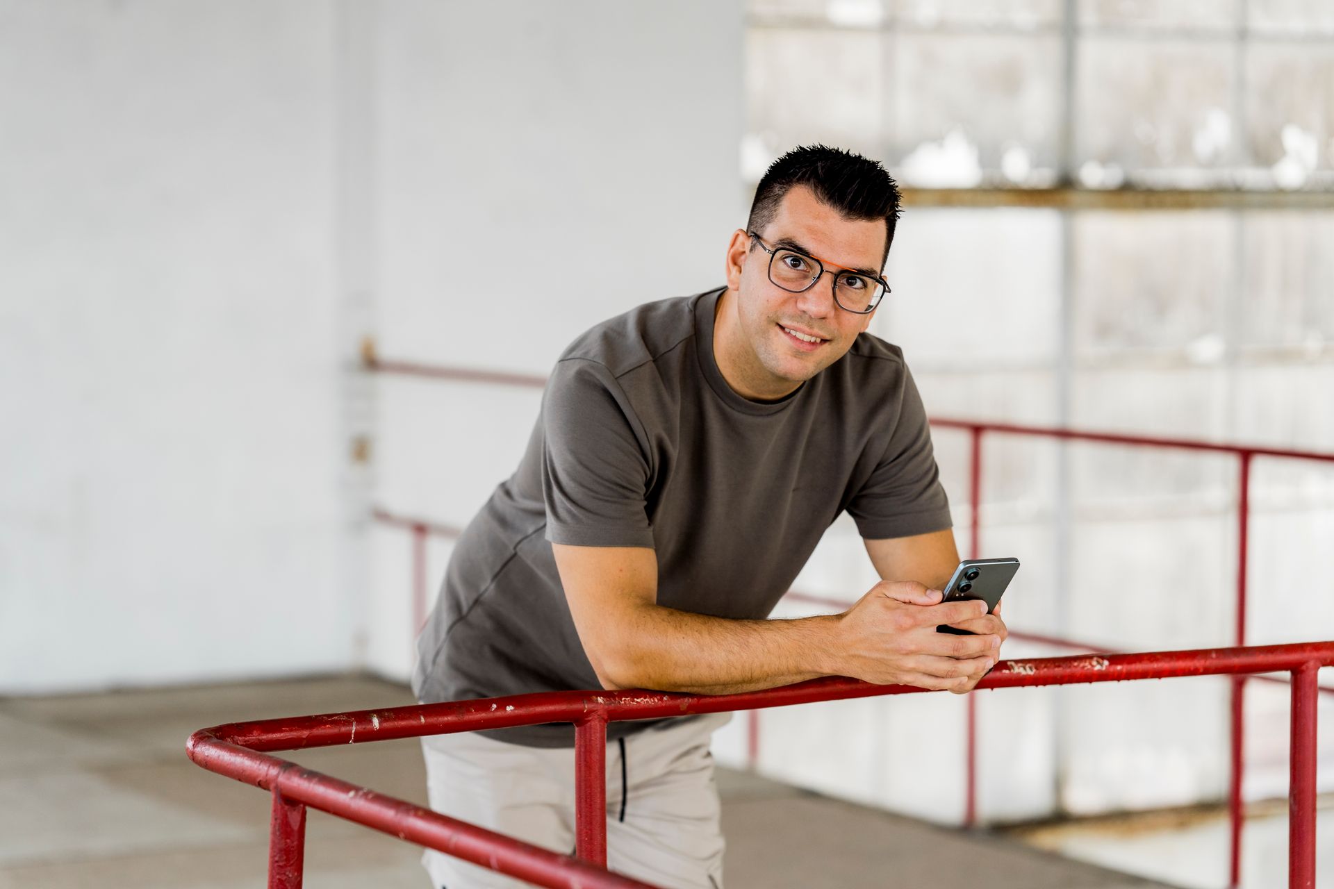 Een man met een bril leunt tegen een rode reling, houdt een telefoon vast en glimlacht.
