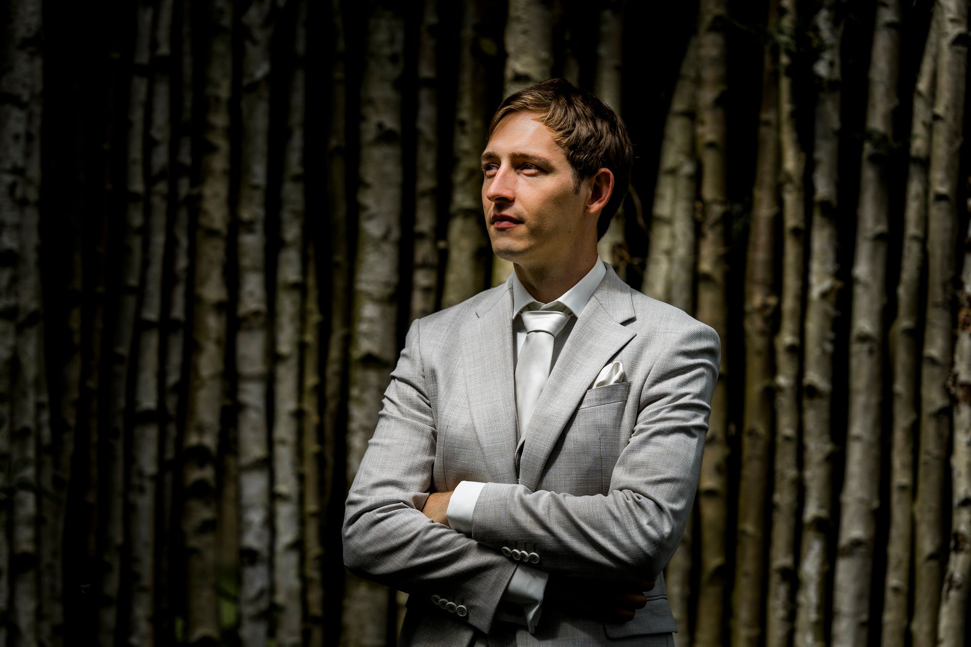 Een man in een grijs pak met de armen over elkaar, staat voor een muur van berkenstammen.