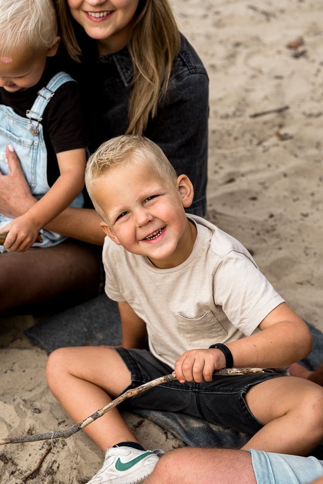 Een jongen glimlacht terwijl hij met een stok in zijn hand in het zand zit, met een gedeeltelijk zichtbare persoon en een kind achter hem.