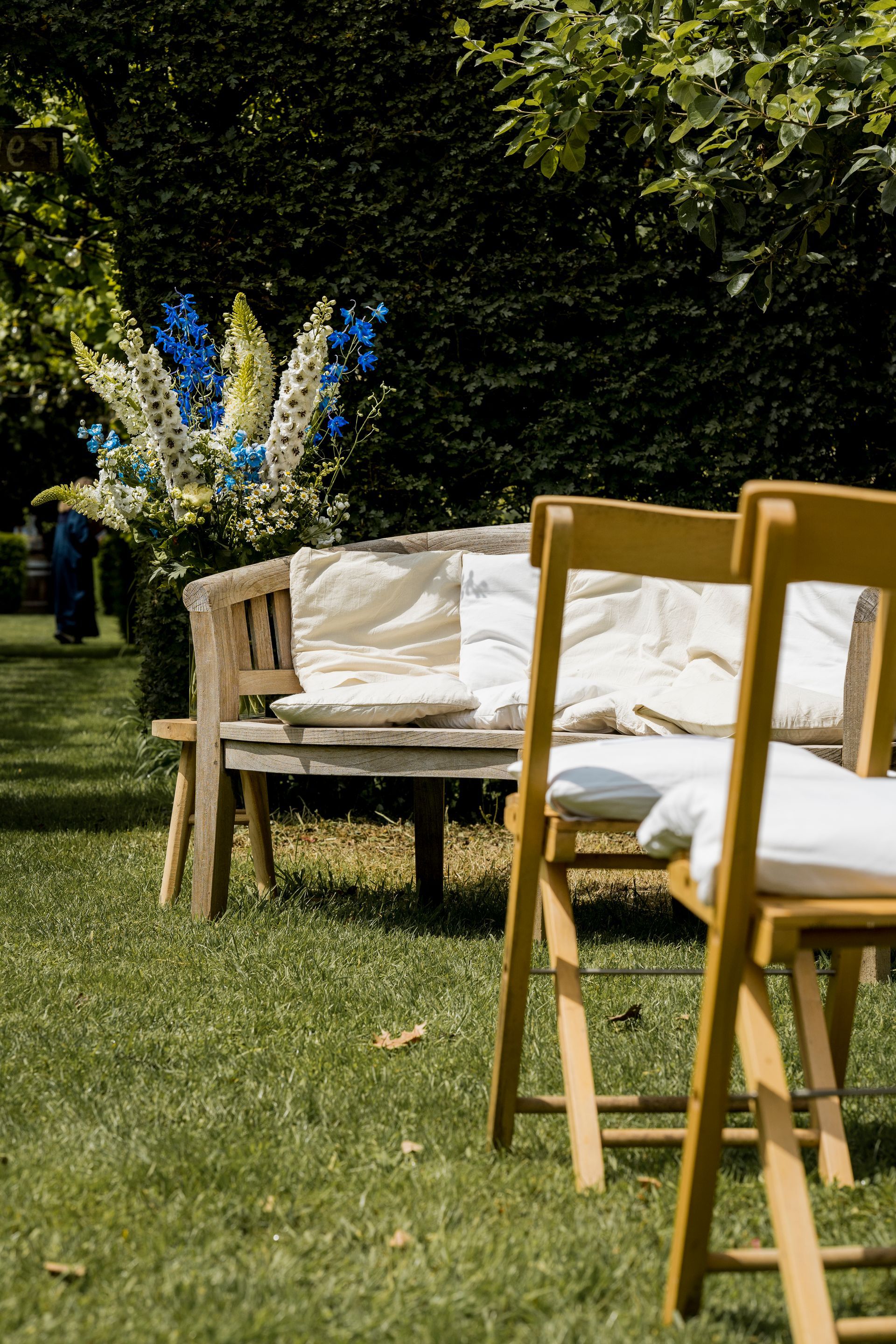 Houten bank met kussens, twee stoelen en een bloemstuk in de tuin.