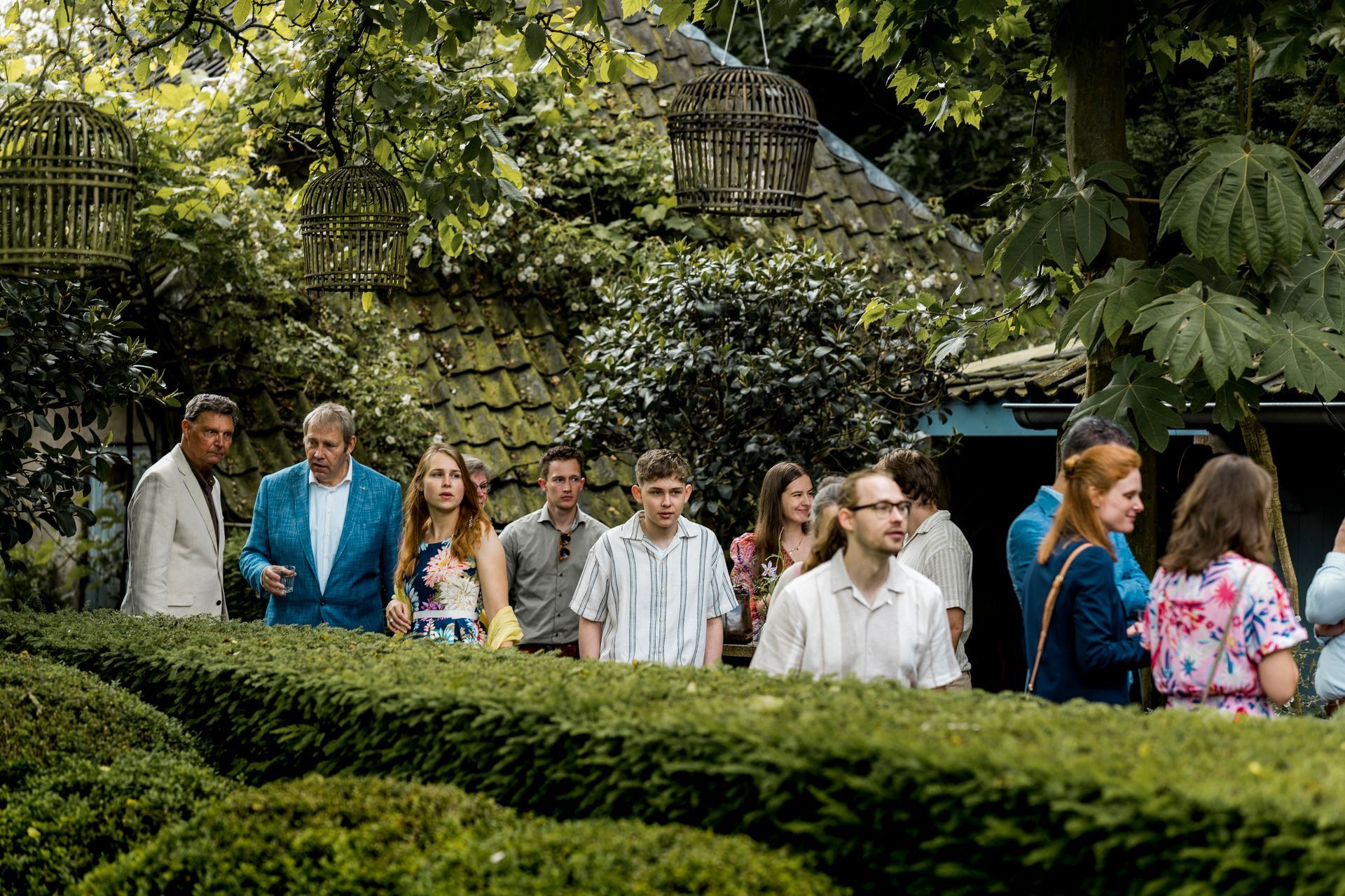 Een groep mensen staat bij een heg in een tuin. Sommigen dragen kleding met bloemenprints.