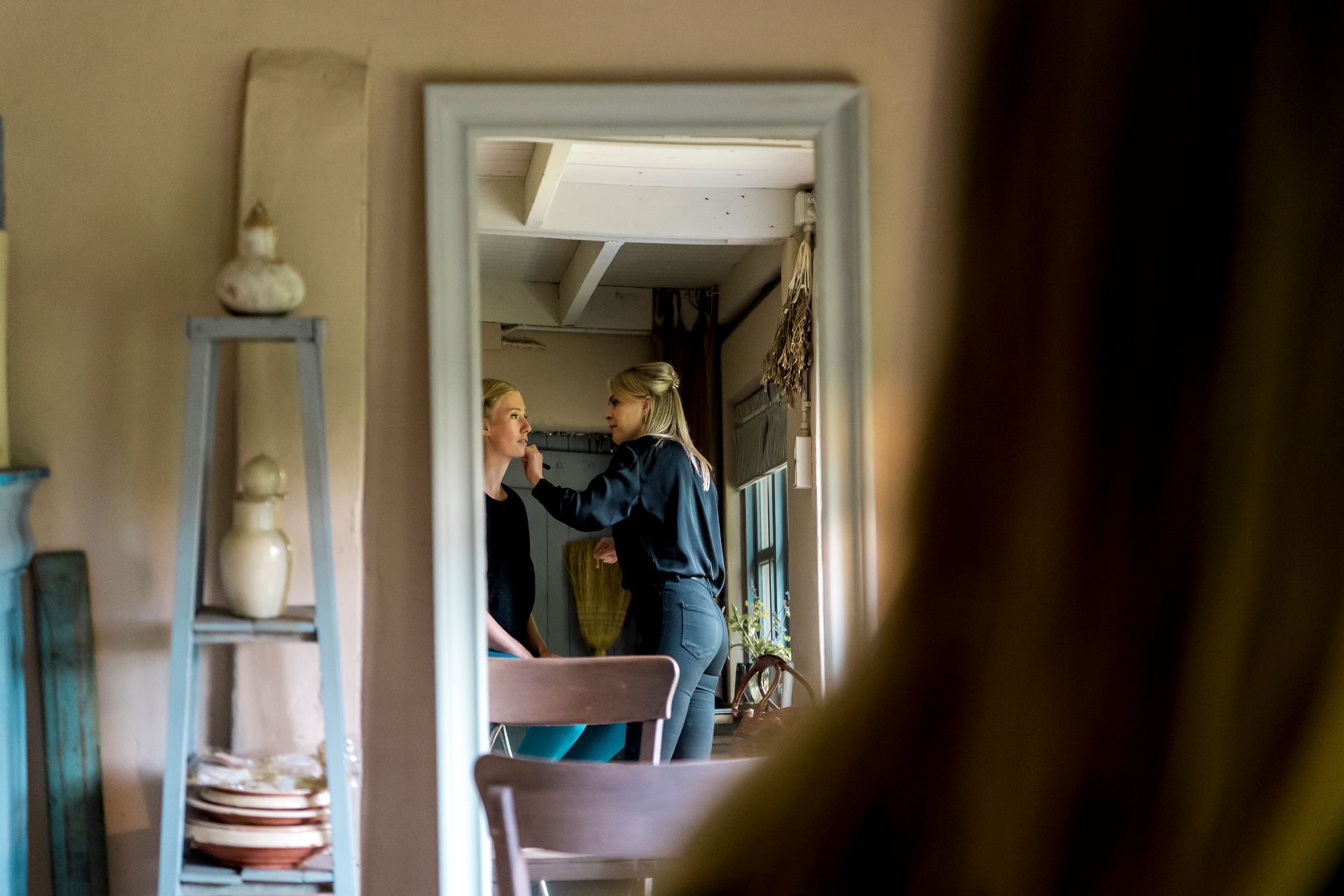 Vrouw brengt make-up aan bij een andere vrouw in een spiegel. Lichtgekleurde kamer, houten plank met aardewerk.