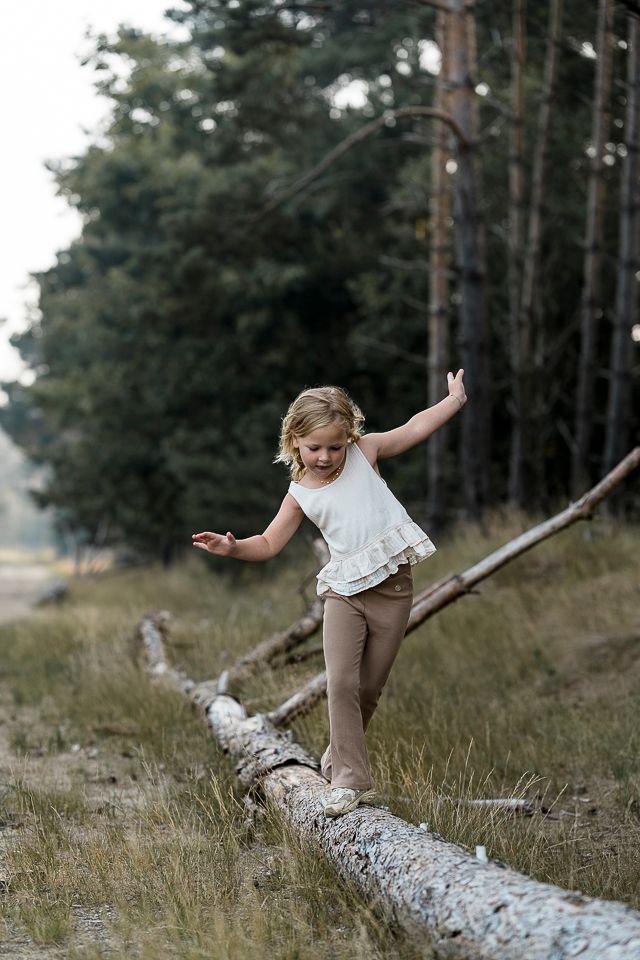 Meisje balanceert op een omgevallen boomstam in een bos, met uitgestrekte armen, bruine broek en witte top.
