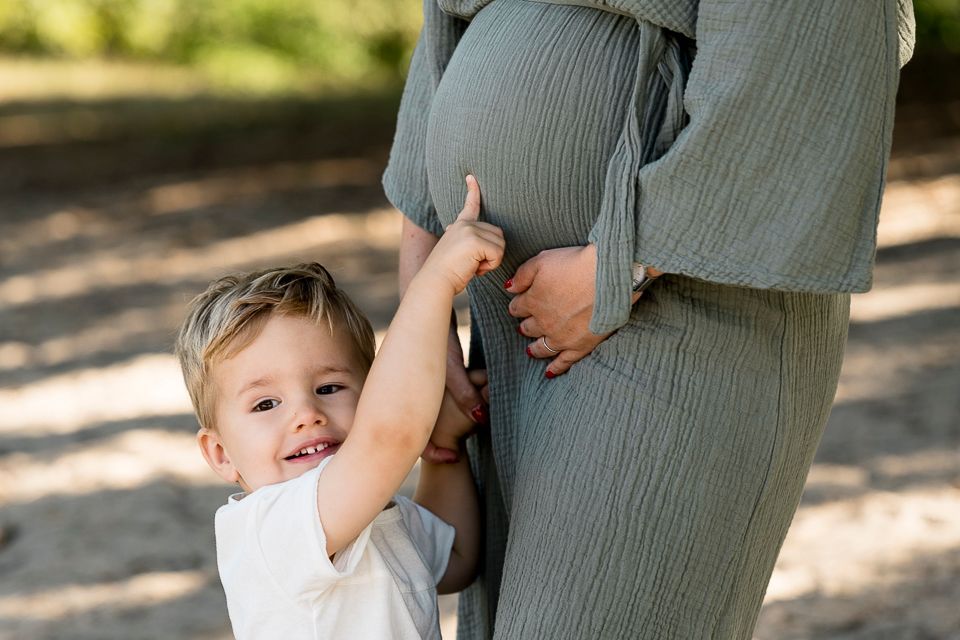 Een jongen wijst naar de buik van een zwangere vrouw; buiten. De zwangere vrouw draagt een groene jurk.